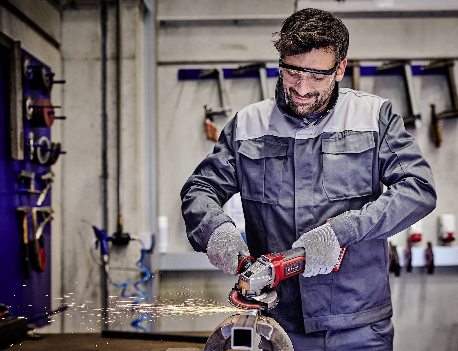 Un homme en tenue de travail meule du métal avec un meuleuse d'angle dans un atelier. Des étincelles volent. Il porte des lunettes de protection et des gants.