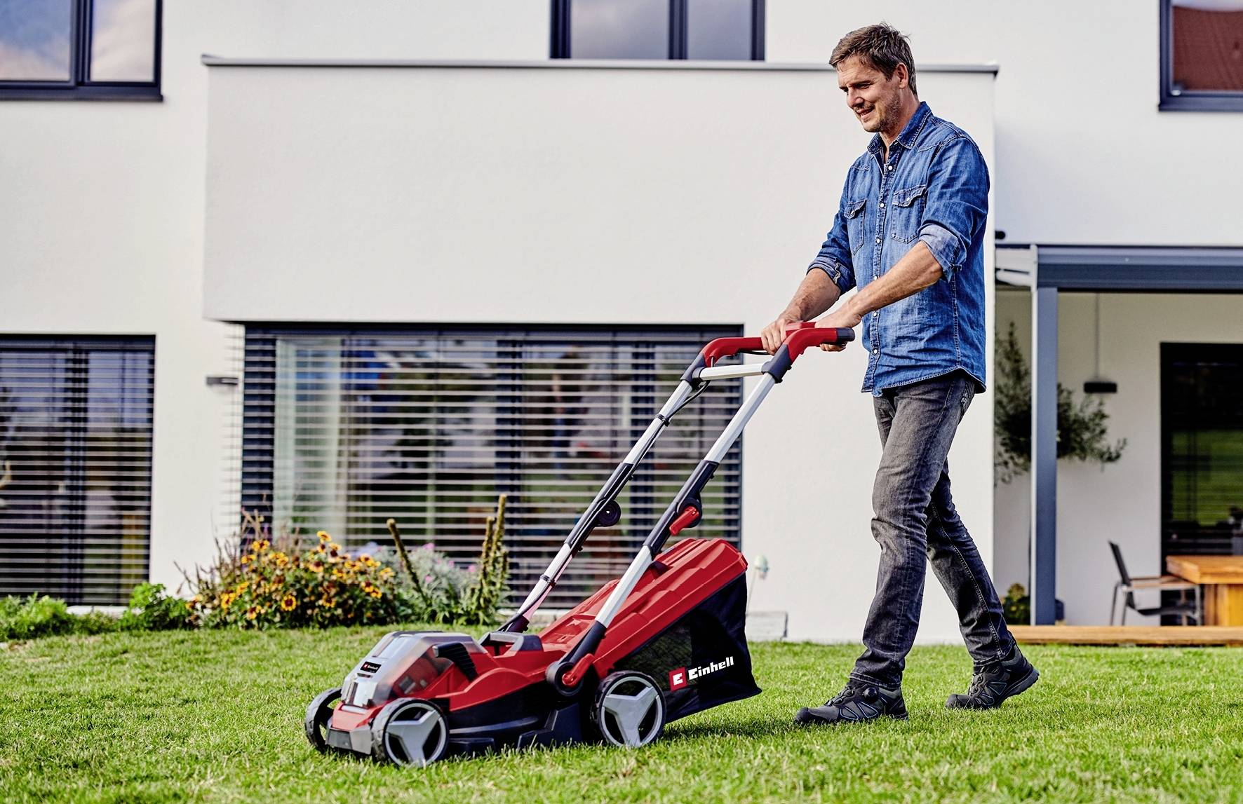 Un homme tond la pelouse devant une maison moderne avec une tondeuse rouge.