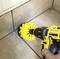 Une main tient un tournevis sans fil jaune équipé d'une brosse rotative qui nettoie les joints de carrelage. Des carreaux sont visibles en arrière-plan.