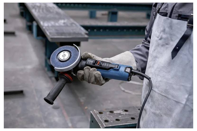 Une personne portant des gants et un tablier tient une meuleuse d'angle bleue, debout sur un sol industriel avec des poutres métalliques en arrière-plan.