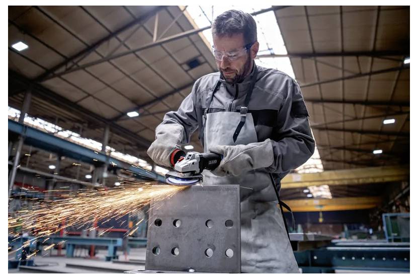 Un homme portant un équipement de protection meule une pièce métallique avec un outil électrique dans un atelier industriel, avec des étincelles qui volent et des machines en arrière-plan.