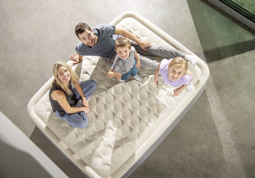 Une famille heureuse de quatre personnes est assise et sourit sur un matelas gonflable dans une pièce moderne.