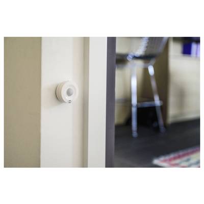 Petit capteur de mouvement blanc monté sur un mur près d'une porte, avec une pièce floue et une chaise en arrière-plan.