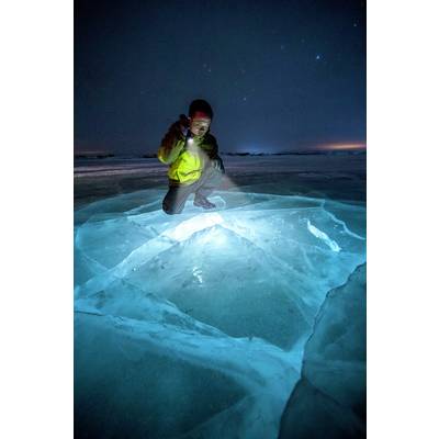 Une personne vêtue d'habits d'hiver est agenouillée sur une surface glacée lumineuse la nuit et l'éclaire avec une lampe de poche pour une investigation.