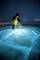 Une personne vêtue d'habits d'hiver est agenouillée sur une surface glacée lumineuse la nuit et l'éclaire avec une lampe de poche pour une investigation.