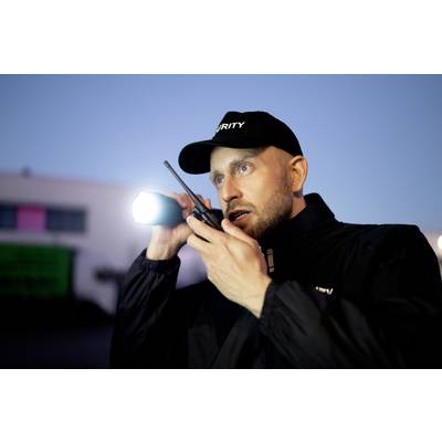 Un agent de sécurité en uniforme se tient dehors la nuit, tenant une lampe de poche et parlant dans une radio.
