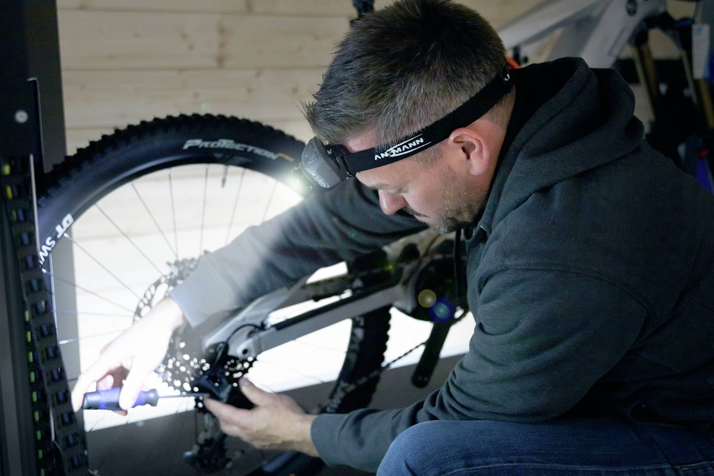 Un homme répare la roue arrière d'un vélo dans un atelier. Il porte un bandeau frontal et utilise un tournevis.