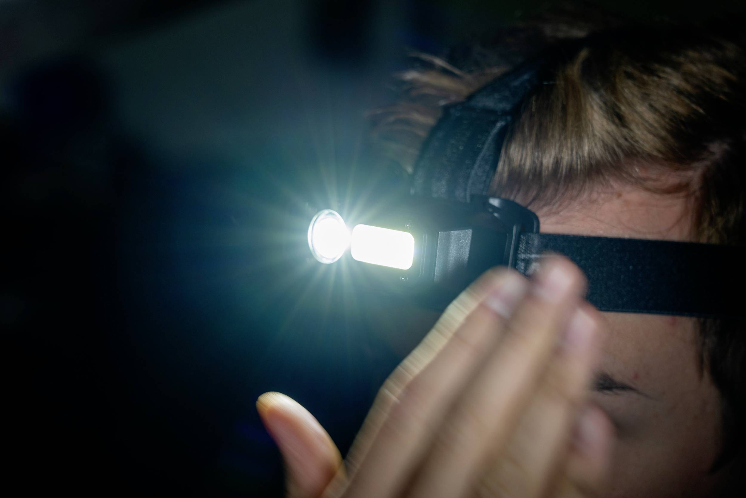 Une personne porte une lampe frontale qui éclaire une pièce sombre. Le faisceau lumineux est intense et la main cache partiellement le visage.