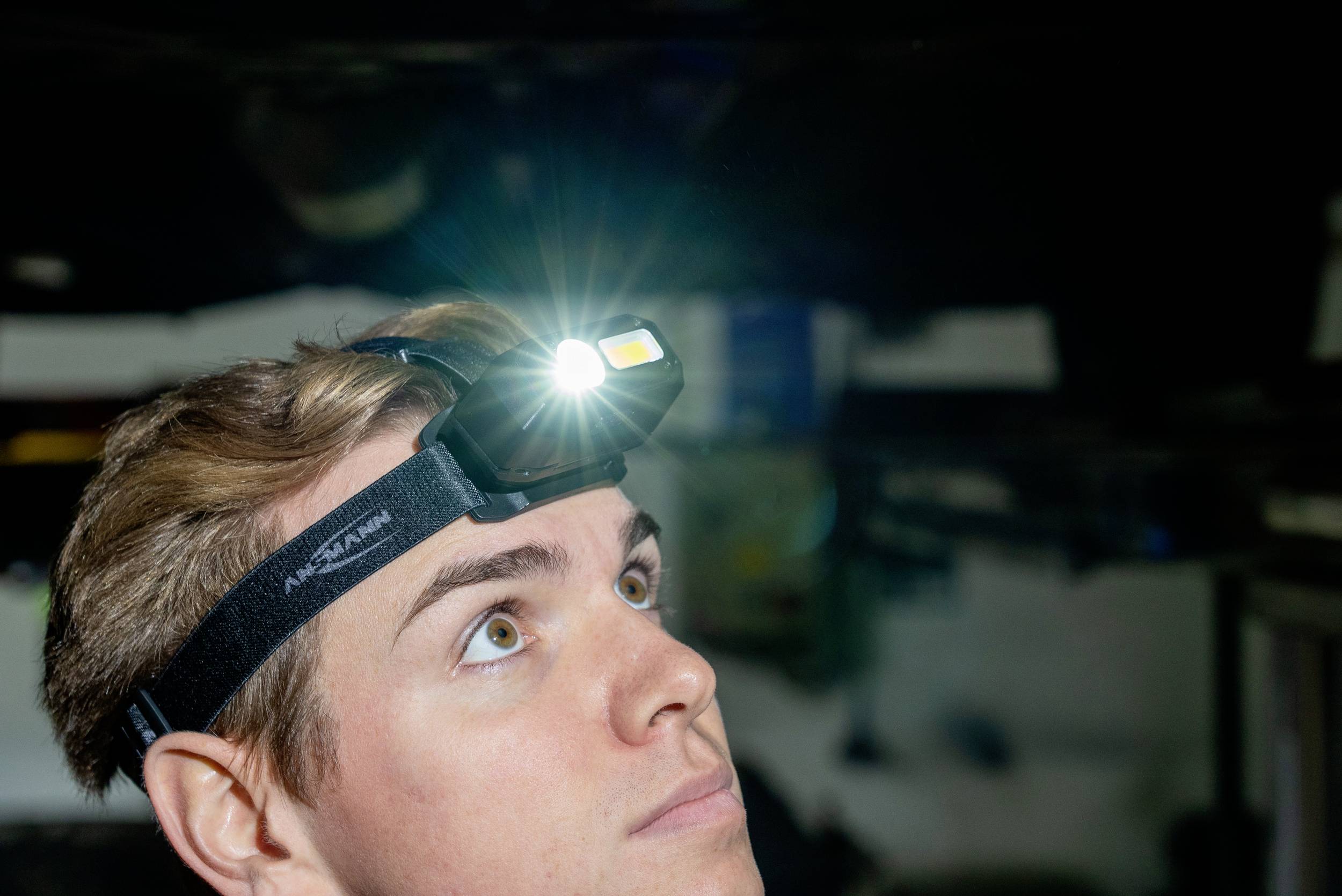 Un homme porte une lampe frontale et regarde vers le haut. La lampe brille intensément, met en valeur son visage et suggère une activité artisanale.