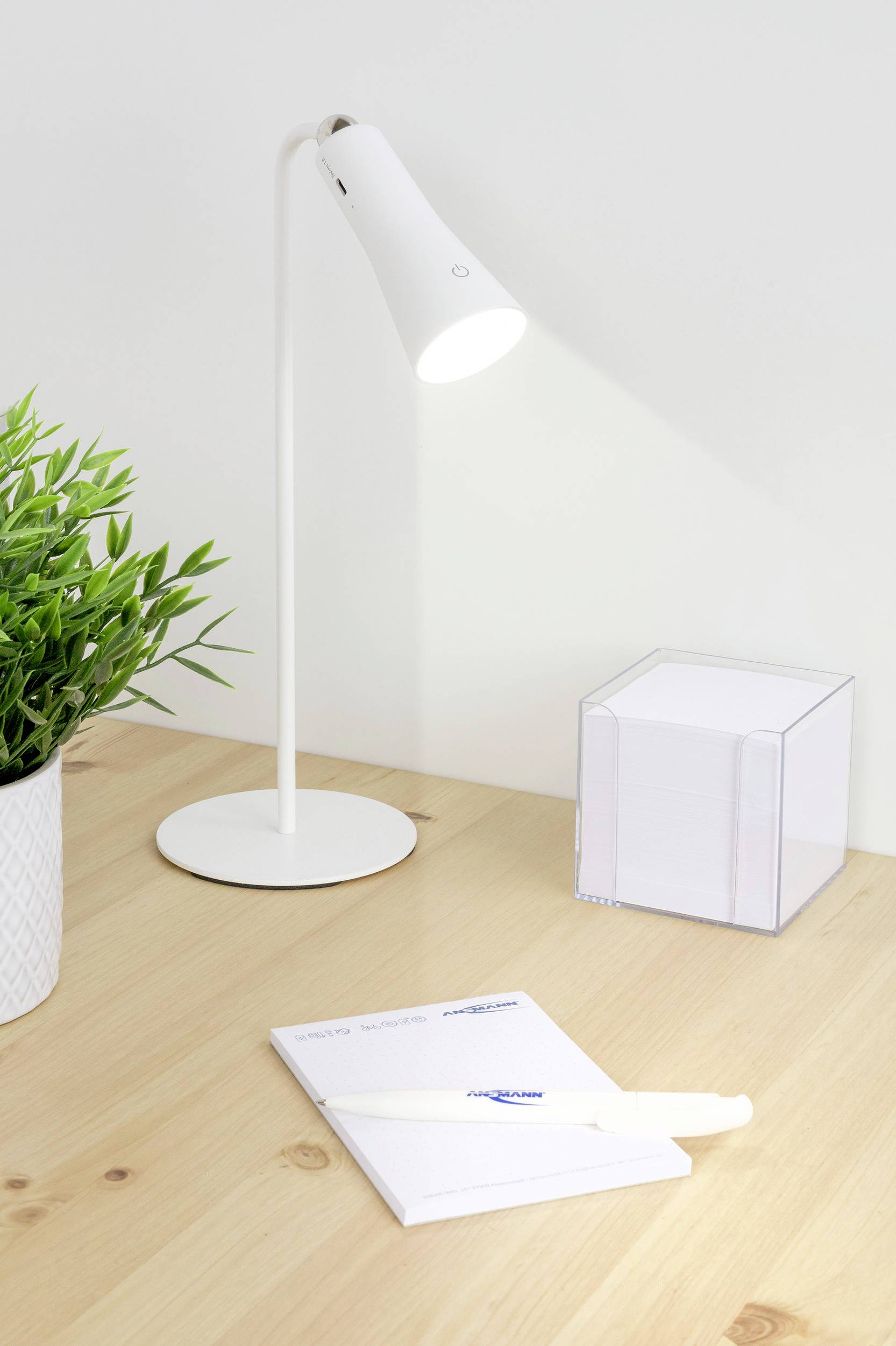 Un bureau minimaliste avec une lampe blanche allumée, un bloc-notes, un stylo et une plante en pot.
