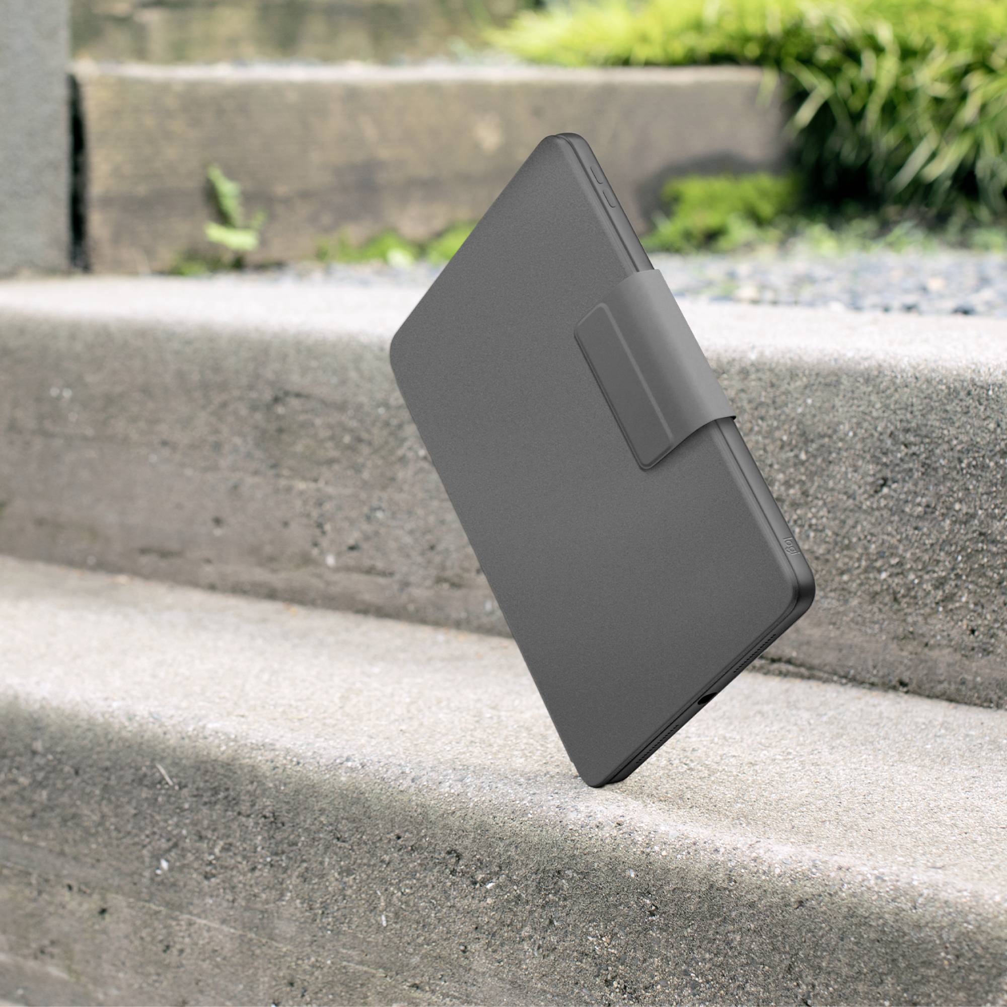 Un lecteur e-book noir et fermé dans un étui de protection est posé en biais sur un escalier en pierre, avec des plantes vertes en arrière-plan.