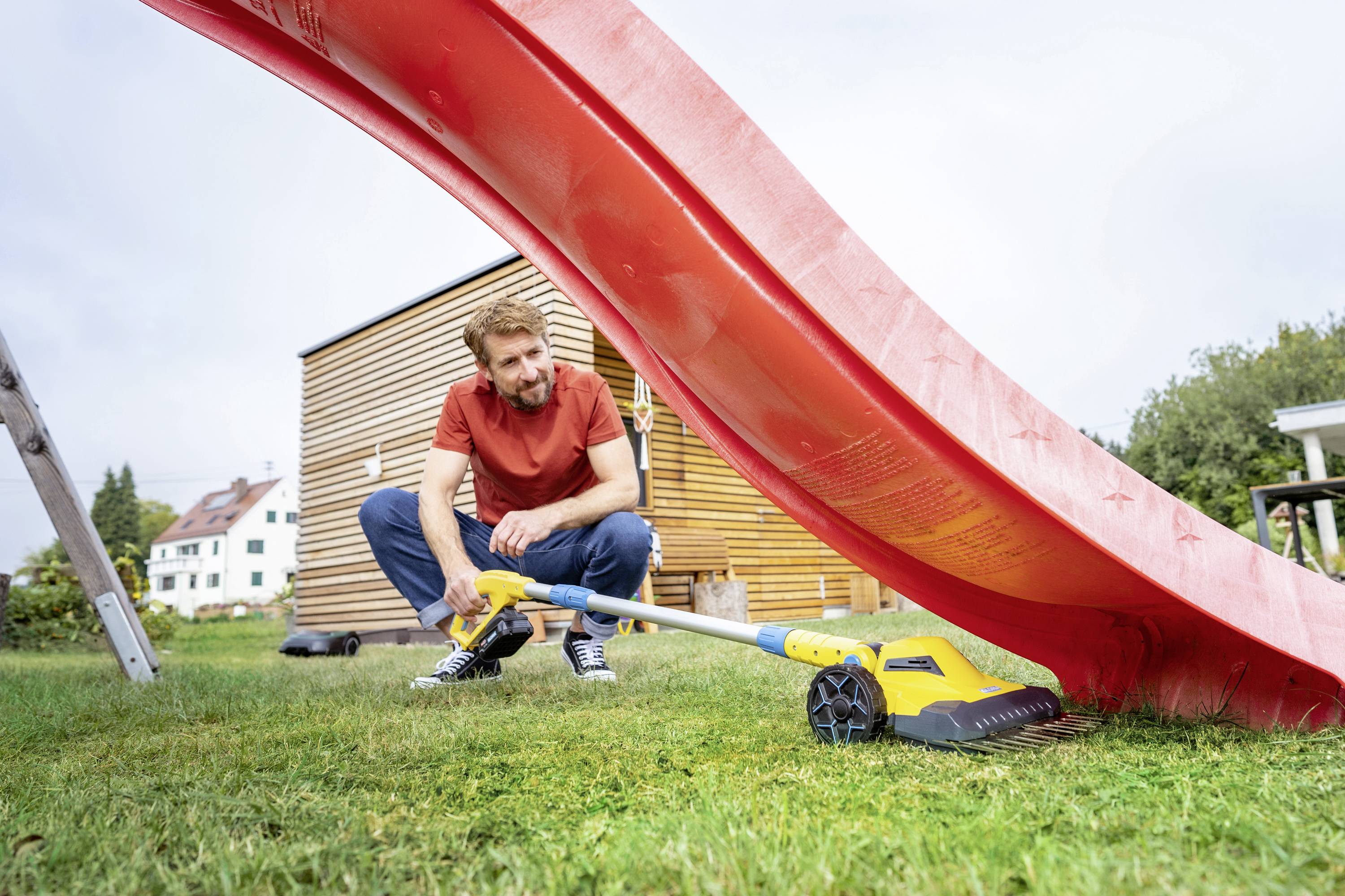 Un homme est à genoux sur la pelouse et nettoie un toboggan rouge à l'aide d'un nettoyeur haute pression. Des maisons sont visibles en arrière-plan.