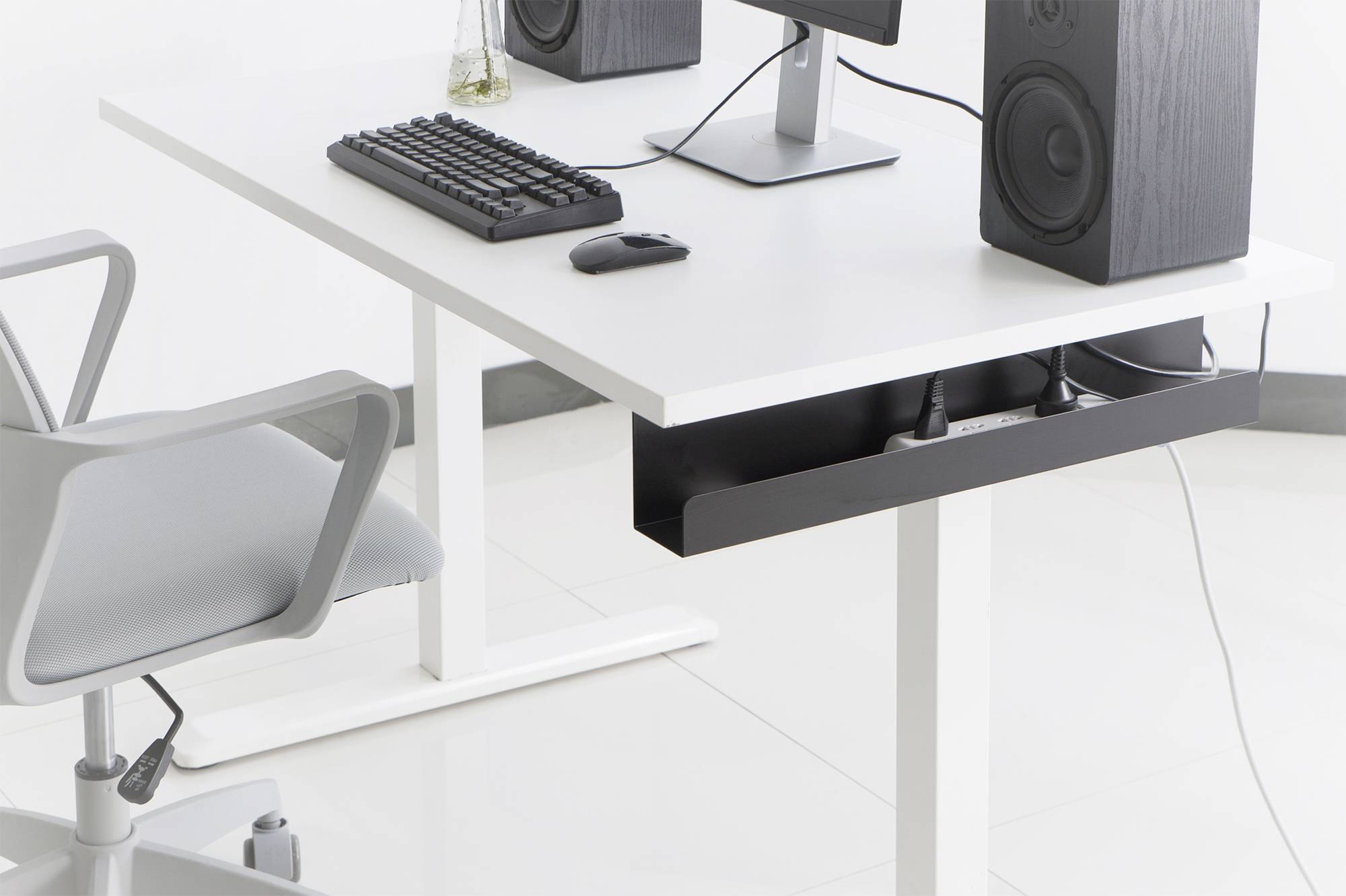 Un bureau blanc avec un écran d'ordinateur, un clavier et des haut-parleurs. Une chaise de bureau grise est placée à côté.