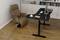 Bureau avec un fauteuil marron et un bureau noir, sur lequel se trouve un écran, un clavier, des plantes, un carnet et une tasse. En arrière-plan : une bibliothèque.