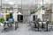 Bureau moderne avec des postes de travail ouverts, des bureaux et des chaises. Des plantes suspendues au plafond. Environnement lumineux et accueillant.