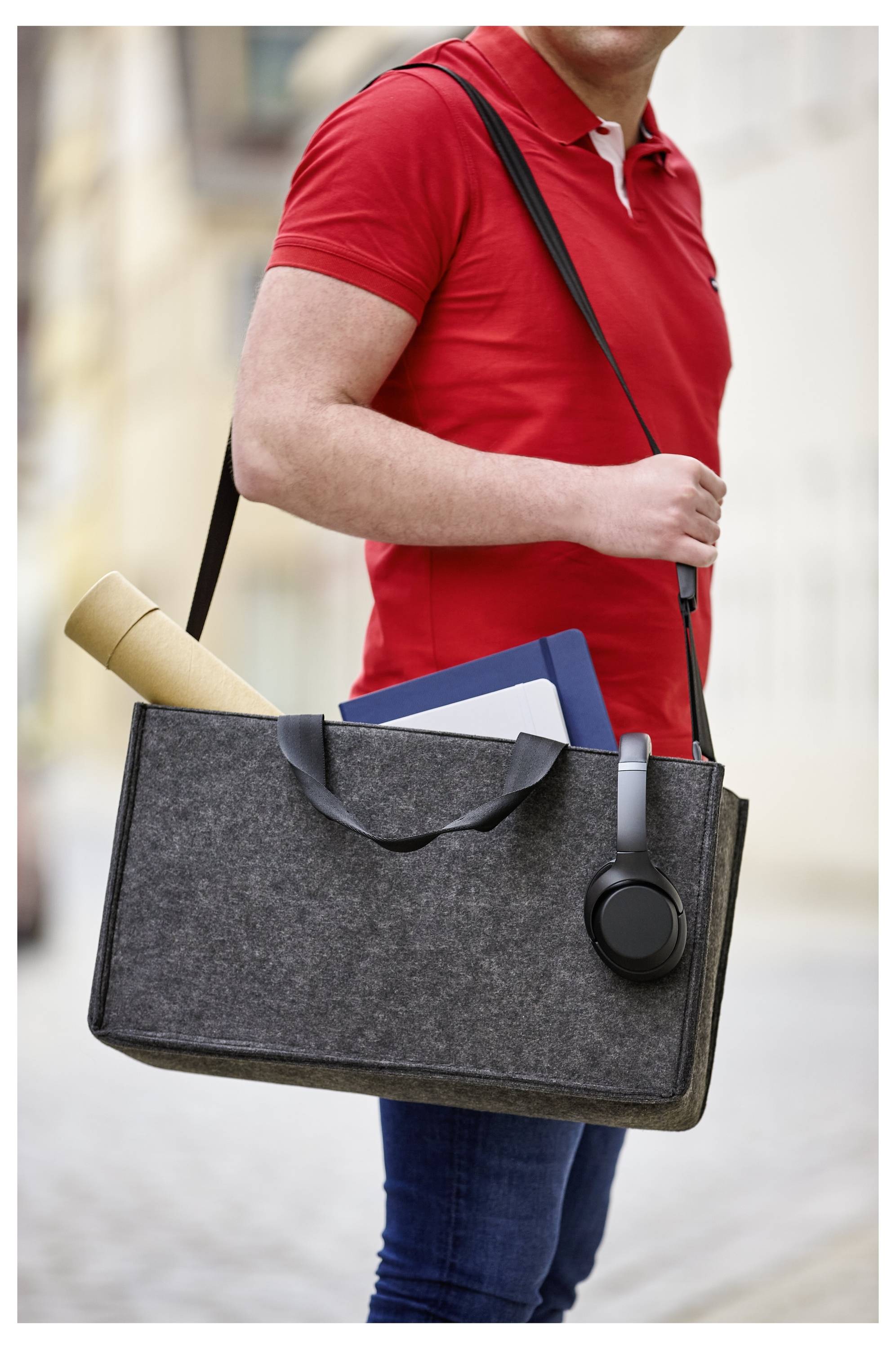 Une personne vêtue d'une chemise rouge portant un grand sac à bandoulière gris rempli de documents et d'un papier enroulé.