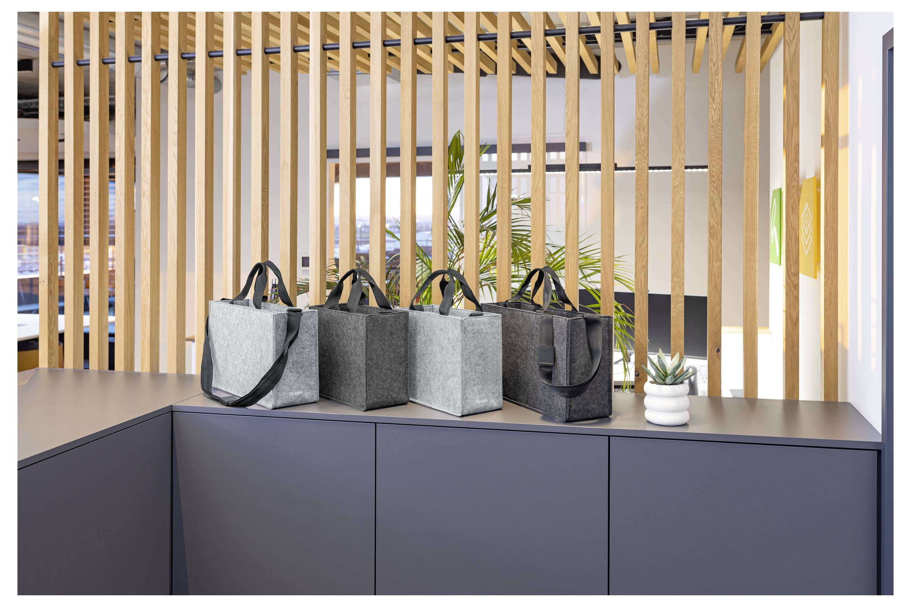 Quatre sacs à main en tissu dans les tons gris et noir sont posés sur un meuble sombre dans un intérieur moderne lumineux avec des lattes de bois verticales et des plantes derrière eux.