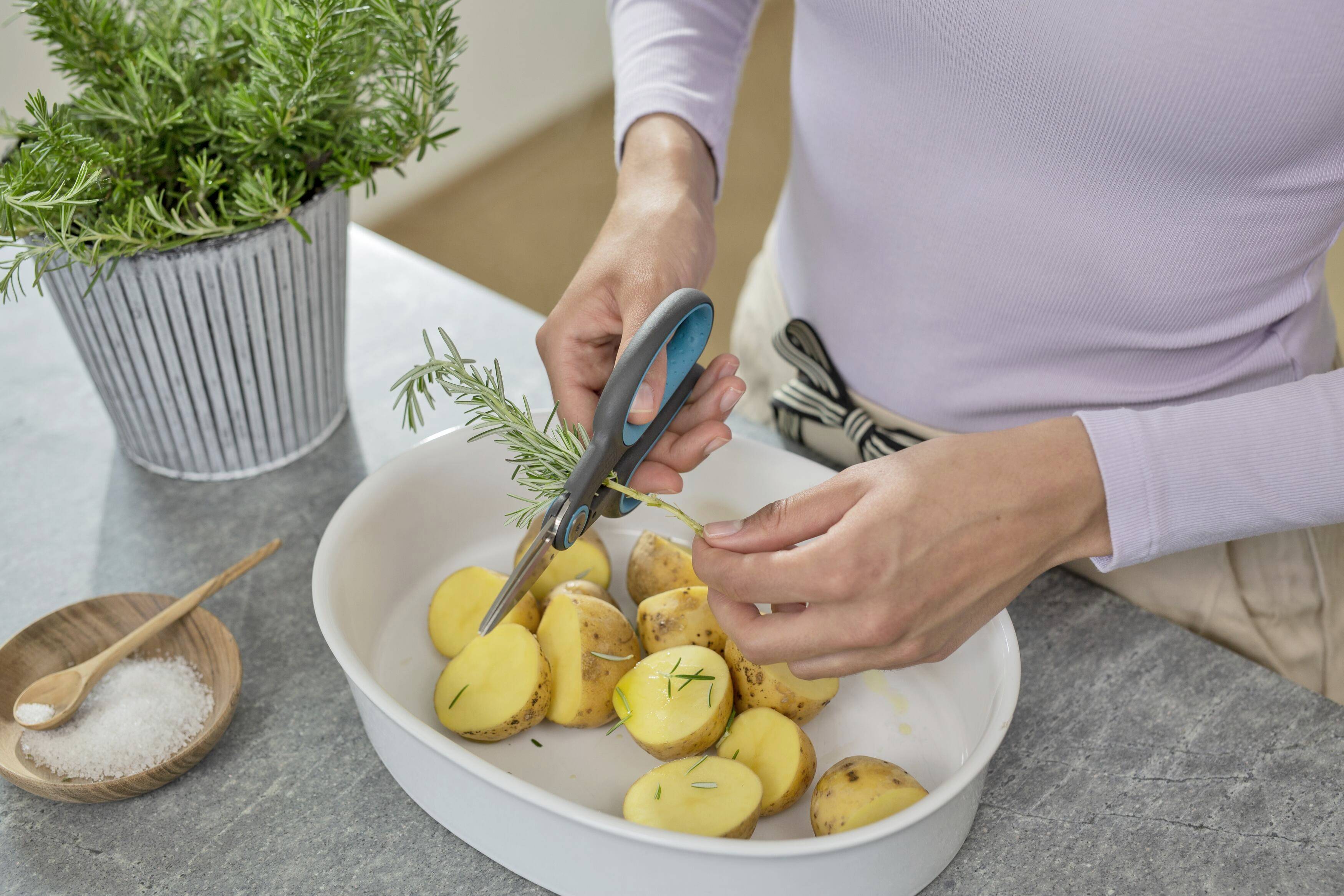 Une femme coupe des herbes fraîches avec des ciseaux et les répand sur des pommes de terre coupées dans un plat à gratin blanc. Des plantes sont visibles en arrière-plan.