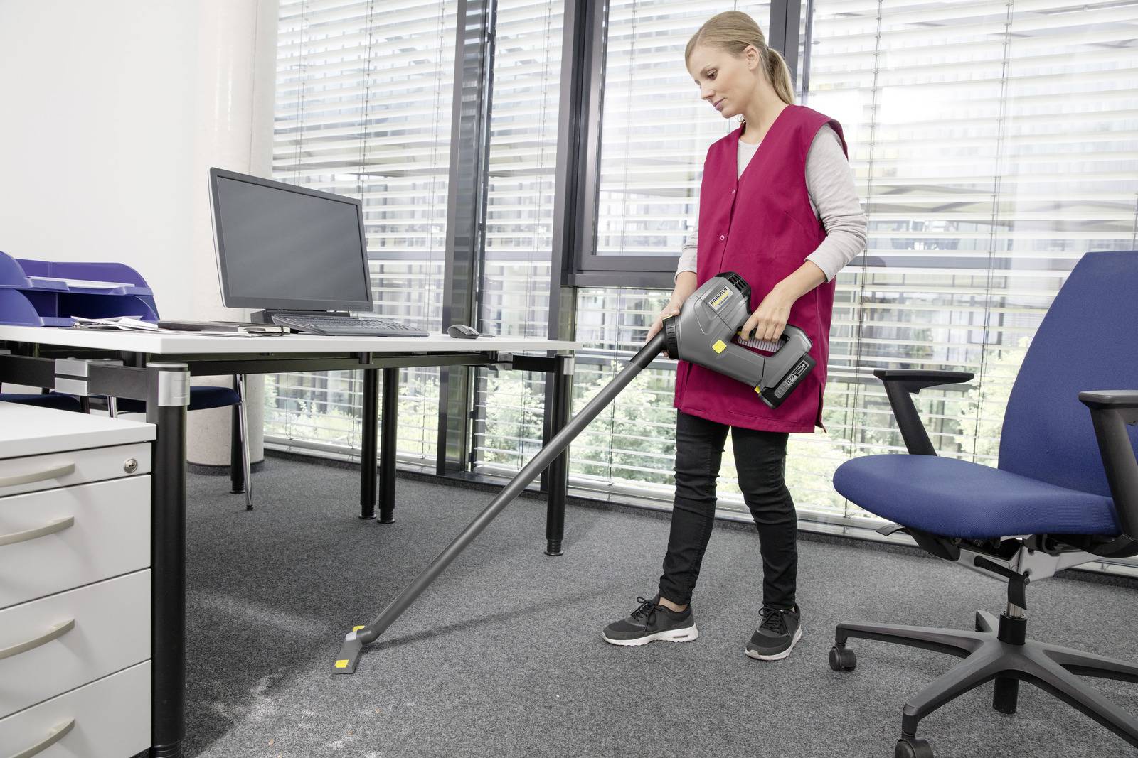 Une personne portant un gilet rose est en train d'aspirer le sol moquetté d'un bureau près d'un bureau avec un écran d'ordinateur, des classeurs et une chaise de bureau.