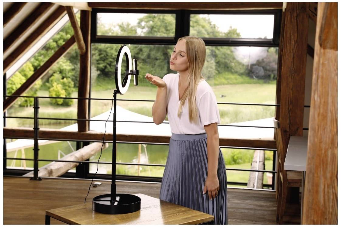 Femme dans un espace lumineux, debout devant un anneau lumineux et une caméra, semblant faire un baiser en direction de l'objectif. Fenêtre en arrière-plan avec une vue verdoyante.