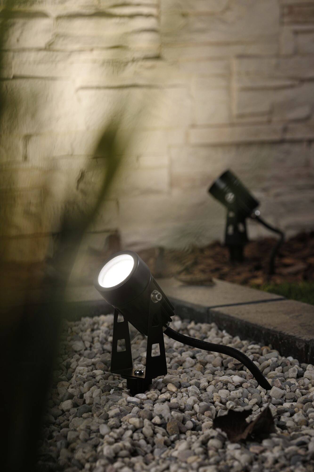 Deux luminaires de jardin éclairent une zone recouverte de pierres blanches au crépuscule, avec un mur de pierre en arrière-plan.