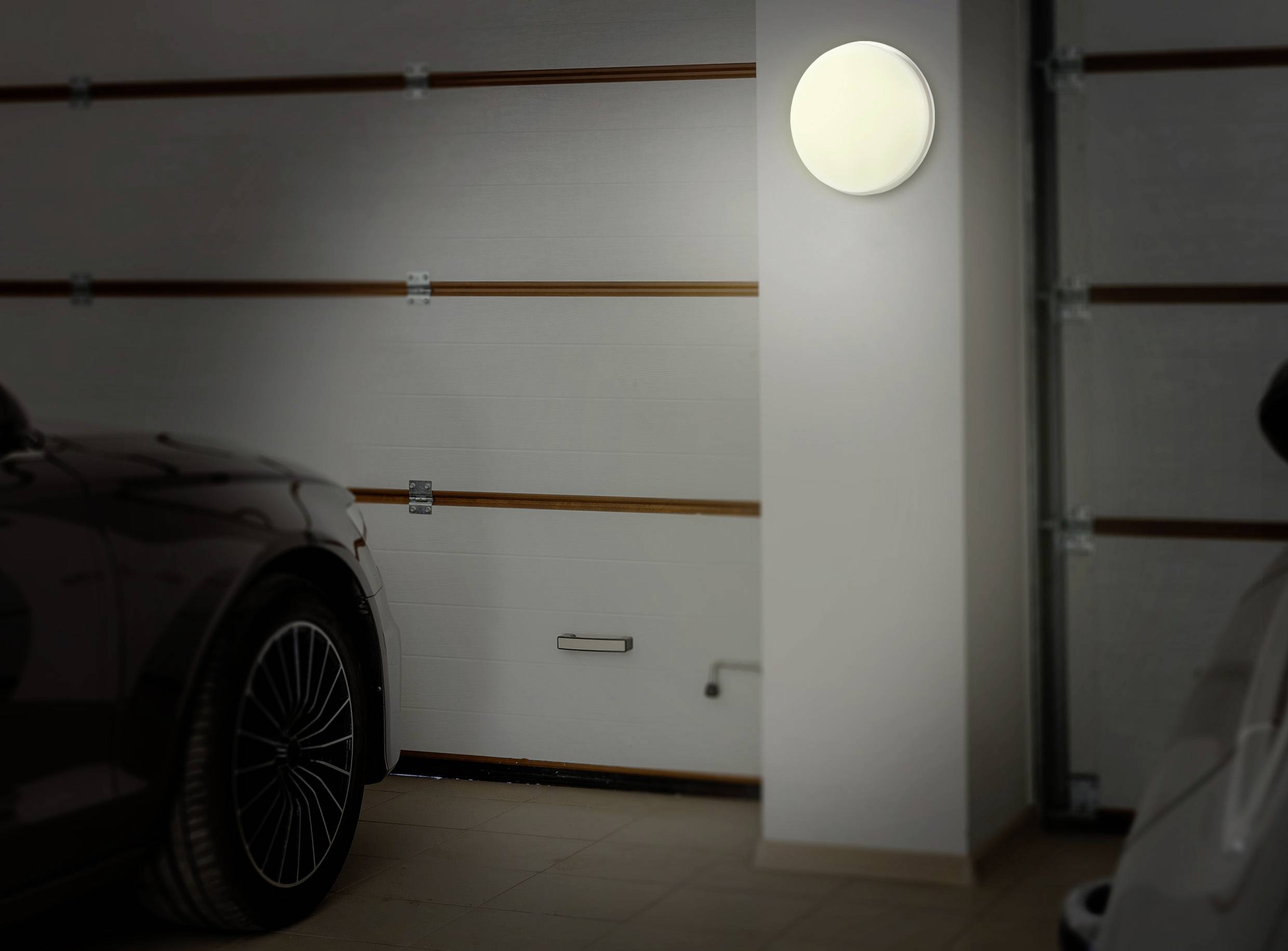 Dans un garage, une voiture est garée à gauche et une autre à droite. Une lampe ronde brille vivement contre le mur blanc.