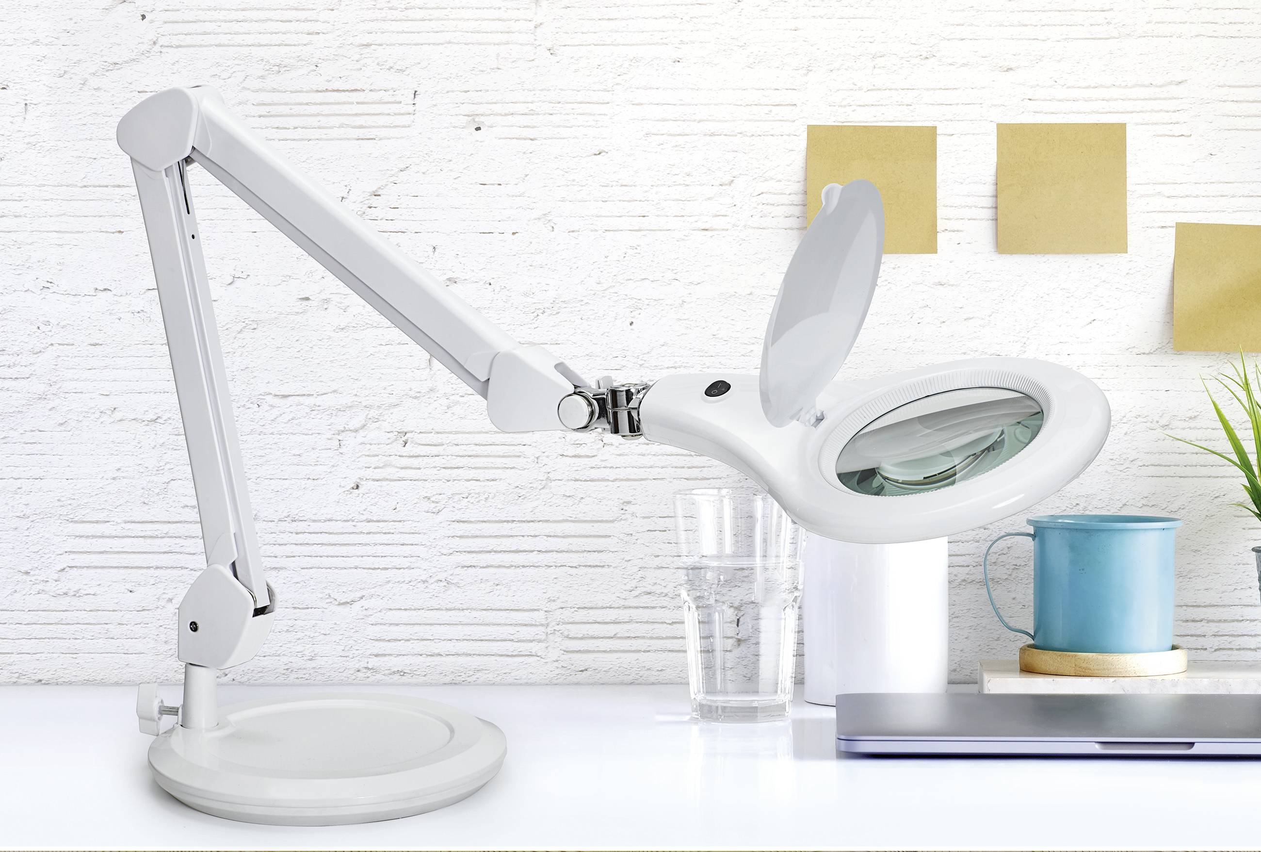 Une lampe de bureau blanche avec une loupe est posée sur une table. En arrière-plan, on aperçoit des notes, un verre, une tasse et un clavier d'ordinateur portable.