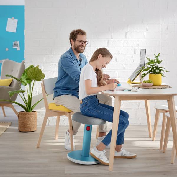 Une femme travaille à une table avec un ordinateur portable, tandis qu'un homme est assis derrière elle et sourit. La pièce est moderne et bien éclairée.