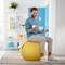 Un homme s'entraîne avec des haltères sur un ballon jaune dans le salon. En arrière-plan, on peut voir une étagère avec des livres et des plantes.