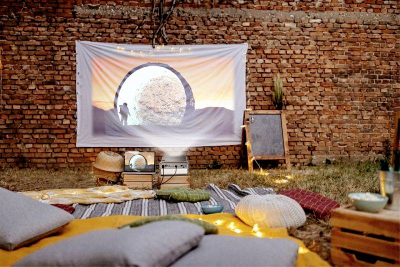Un cinéma en plein air confortable avec des couvertures et des coussins sur le sol, un projecteur diffuse un film sur un drap tendu contre un mur de briques.