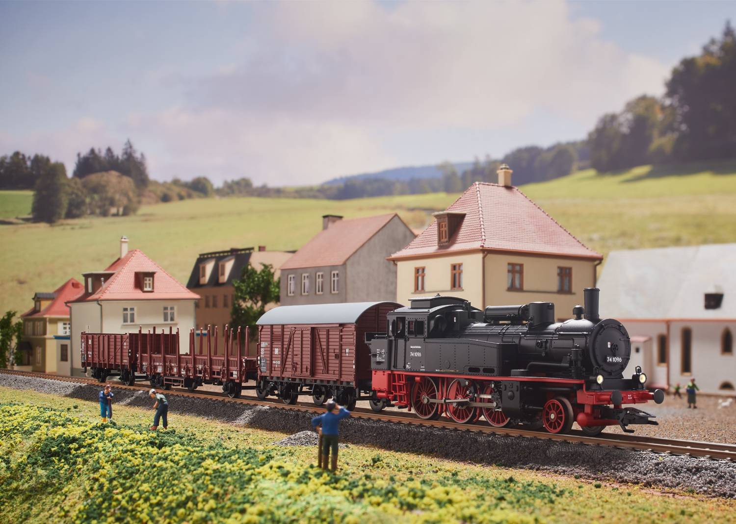 Une locomotive miniature tire des wagons sur un parcours de train miniature à travers un paysage rural avec de petits bâtiments et des figurines.