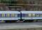 Train de modélisme ferroviaire composé de deux wagons bleus éclairés, roulant sur une voie ferrée avec des bâtiments flous en arrière-plan.