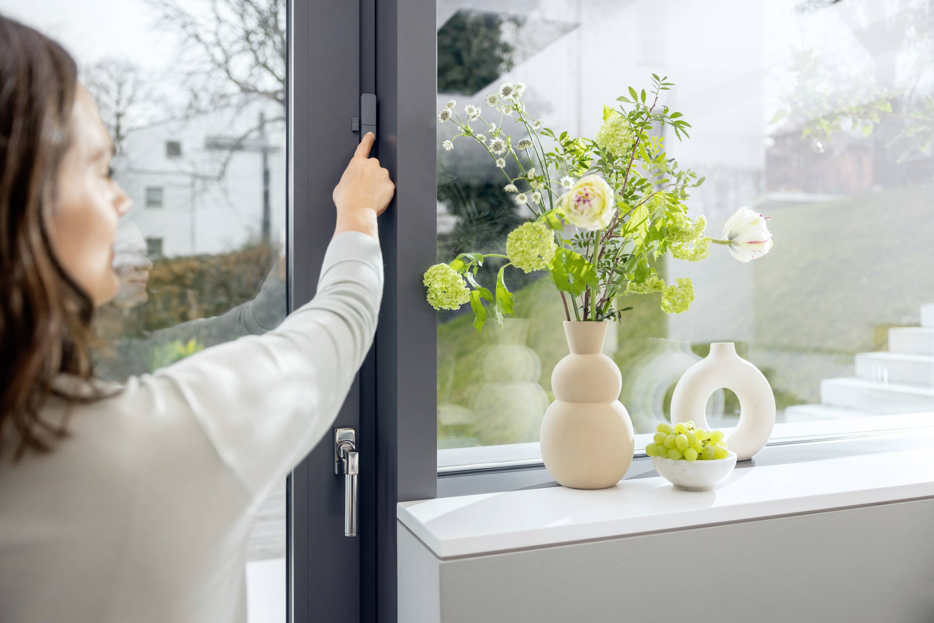 Une femme ouvre une fenêtre à côté d'un rebord de fenêtre garni de vases remplis de fleurs et d'un bol de raisins verts.