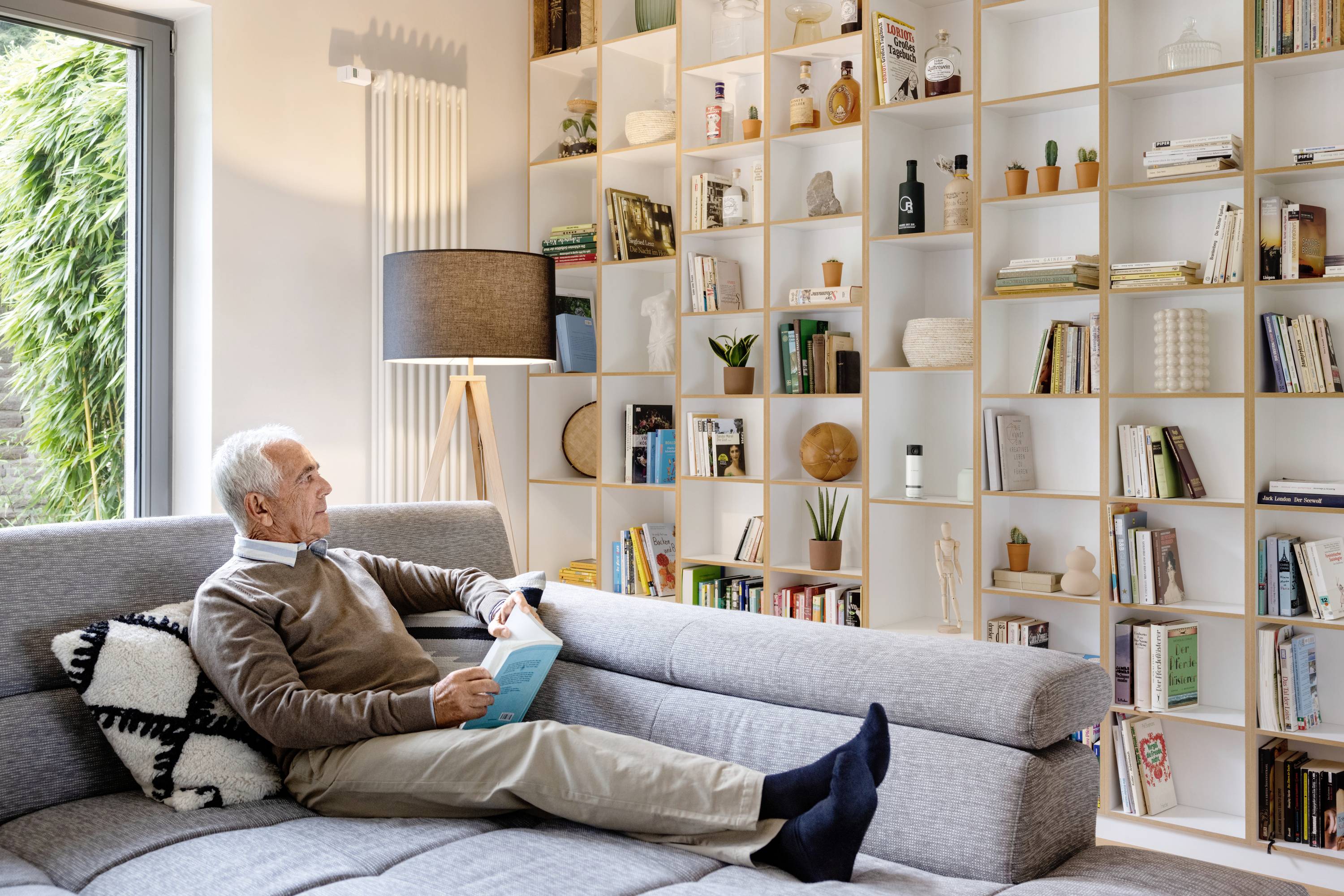 Un homme âgé est assis sur un canapé gris dans un salon moderne et lit un livre. Des étagères à livres sont disposées en arrière-plan.