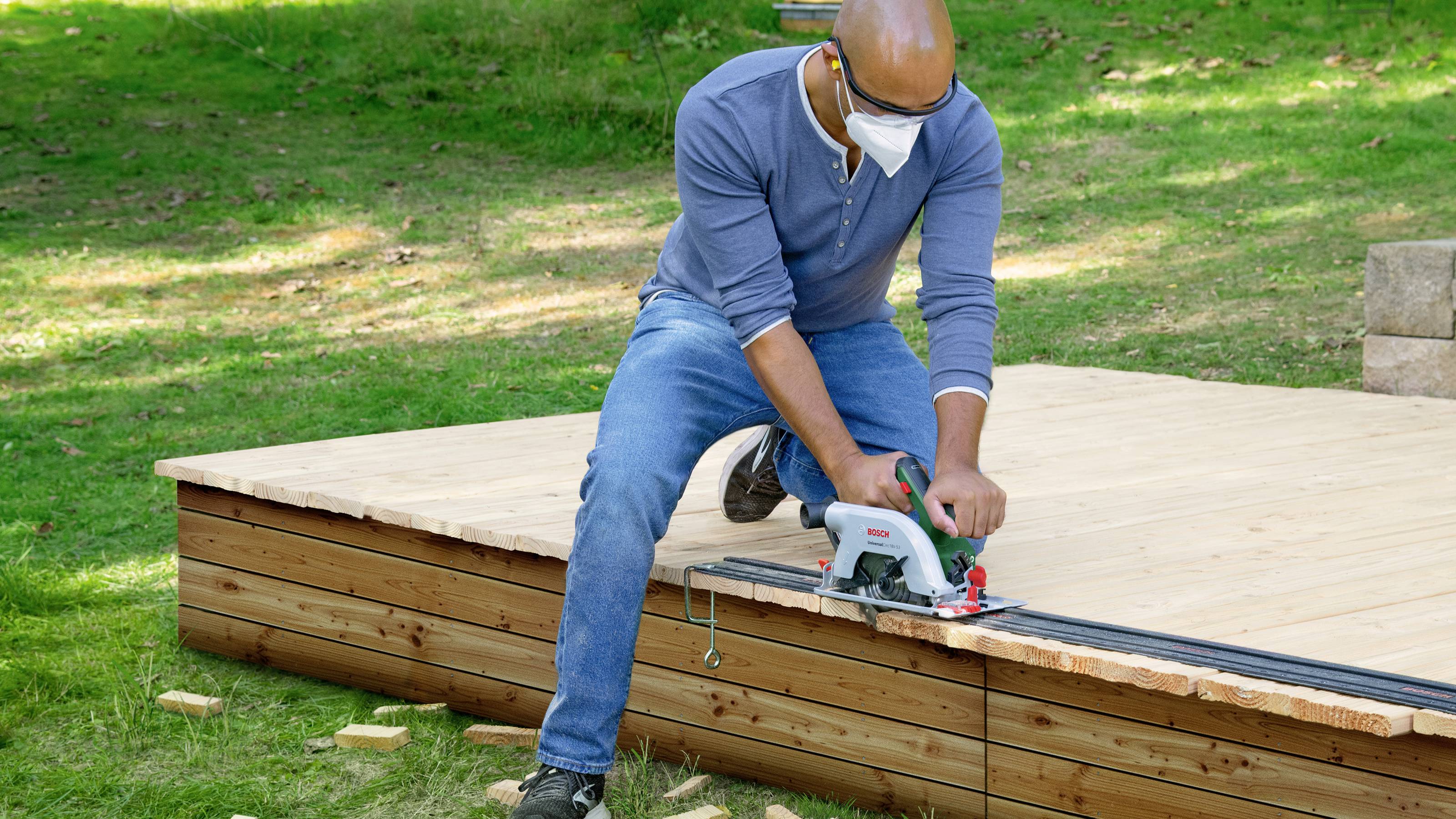 Une personne scie une terrasse en bois dans le jardin avec une scie circulaire. Il porte des lunettes de protection et un masque. De la pelouse environnante et du soleil sont présents.