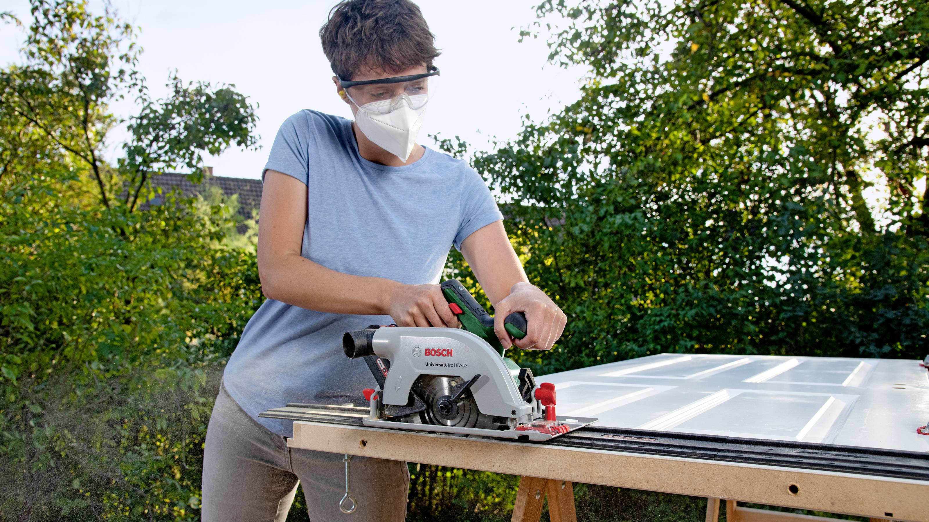Une personne scie une planche à l'extérieur à l'aide d'une scie circulaire portative électrique. Des lunettes de sécurité et un masque sont portés. Des arbres sont visibles en arrière-plan.