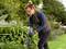 Une personne dans le jardin coupe des plantes avec des ciseaux. Elle porte des lunettes et des gants, entourée de buissons verts et de fleurs.