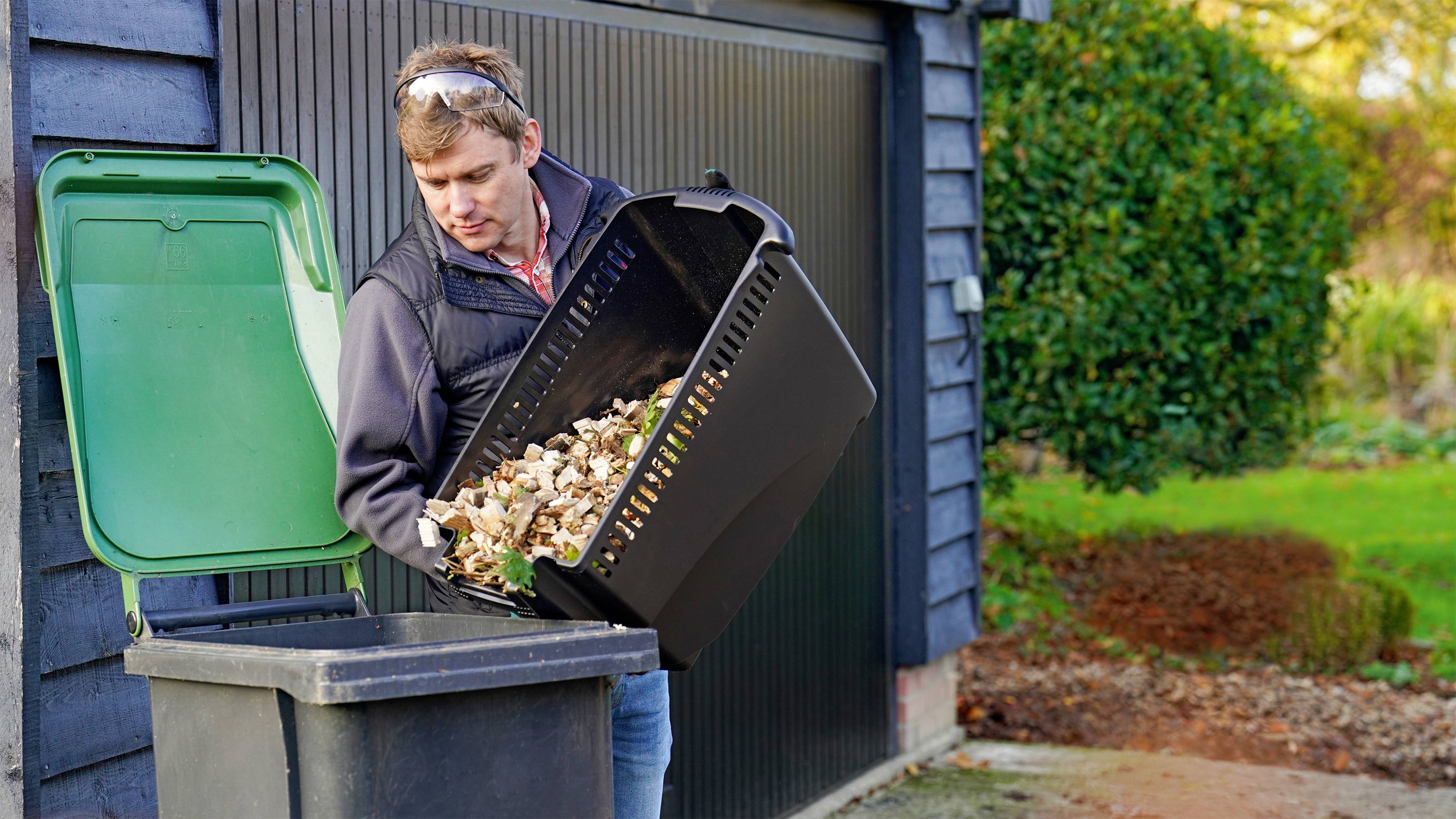 Une personne vide un récipient de déchets de jardin dans une poubelle devant une porte de garage. Des plantes vertes sont visibles en arrière-plan.
