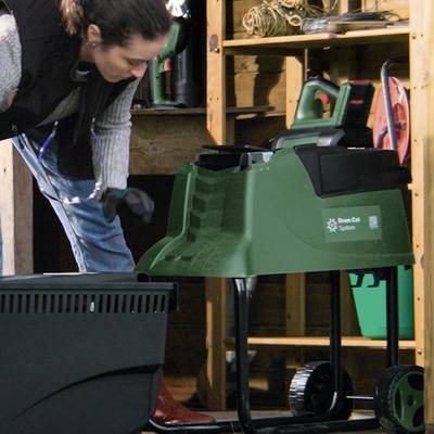 Une personne travaille dans un atelier avec un aspirateur de feuilles vert. Des outils et des étagères en bois sont visibles en arrière-plan.
