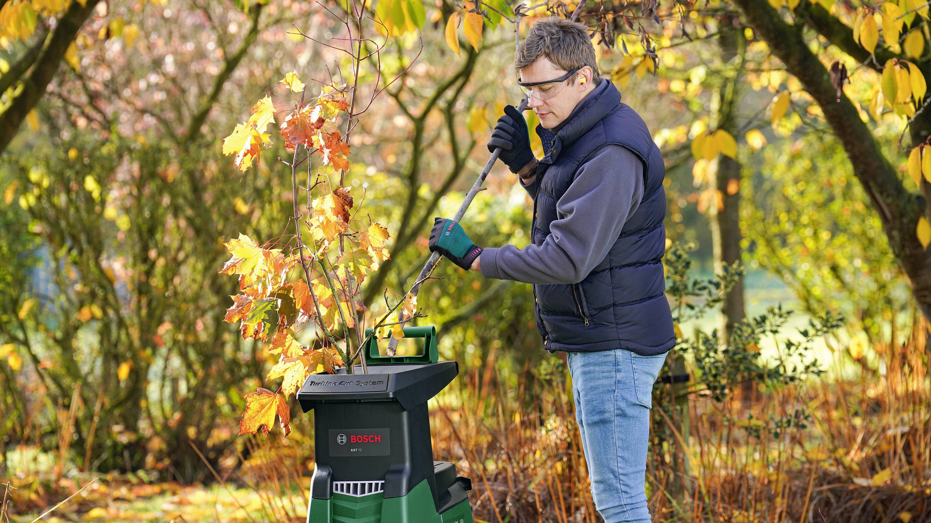 Un homme broie des feuilles avec un broyeur dans un jardin d'automne. Il porte des lunettes de protection et des vêtements chauds.