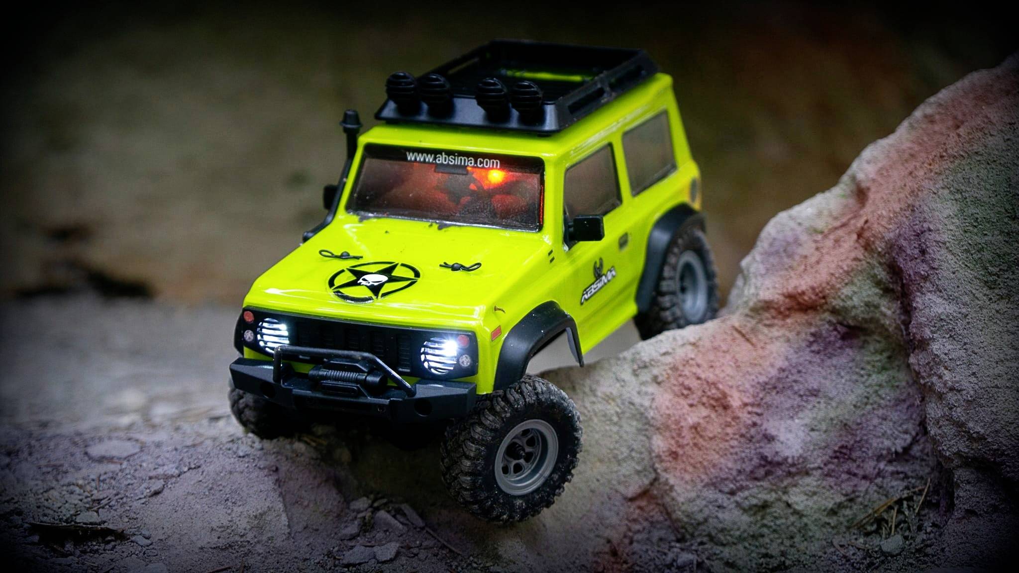 Un véhicule tout-terrain jouet jaune vif escalade un terrain rocailleux. Les phares avant sont allumés, avec des pneus adaptés au terrain.