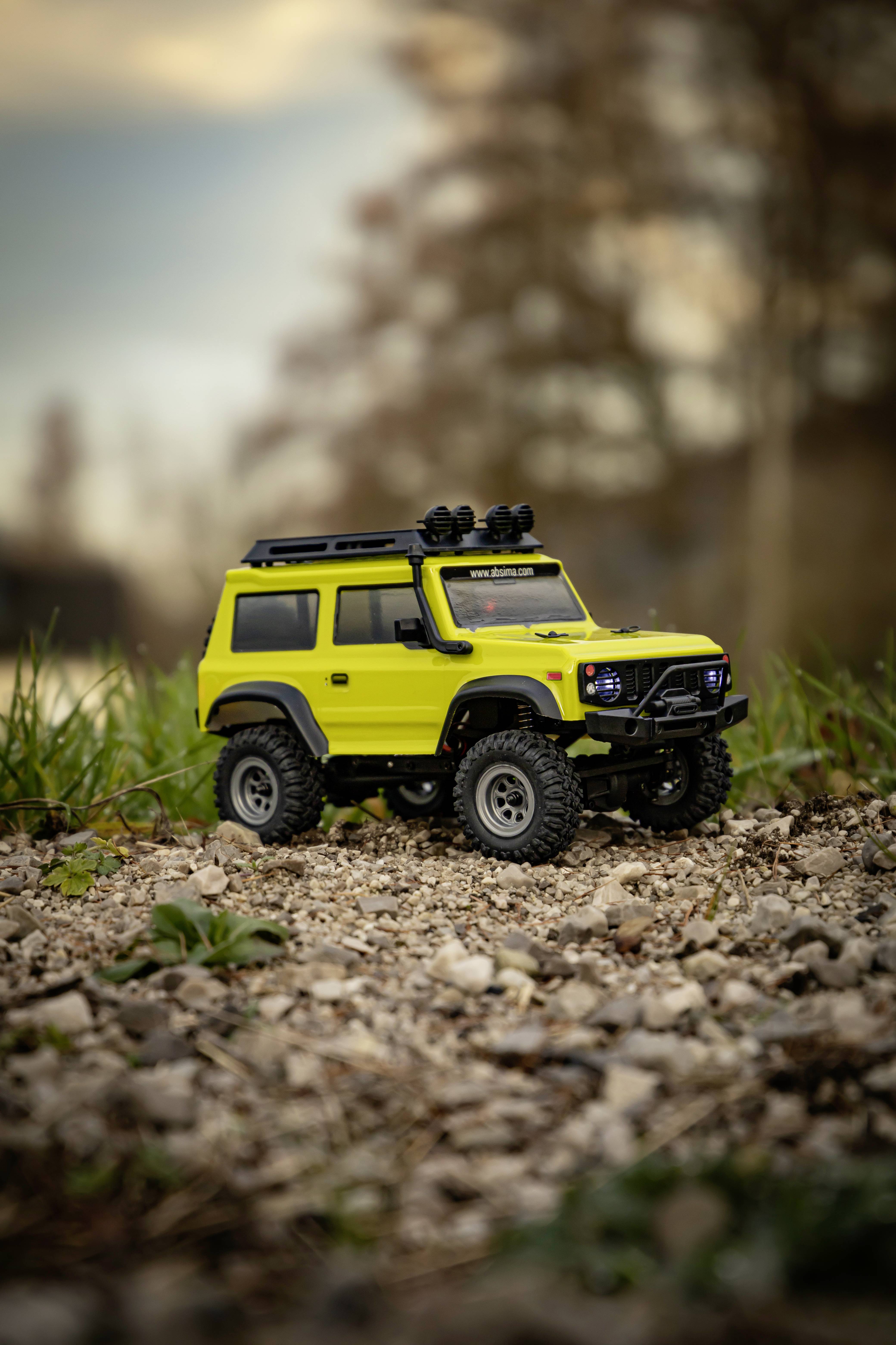 Une voiture miniature jaune de style tout-terrain est posée sur un sol pierreux, entourée d'herbe verte et avec un arrière-plan flou.