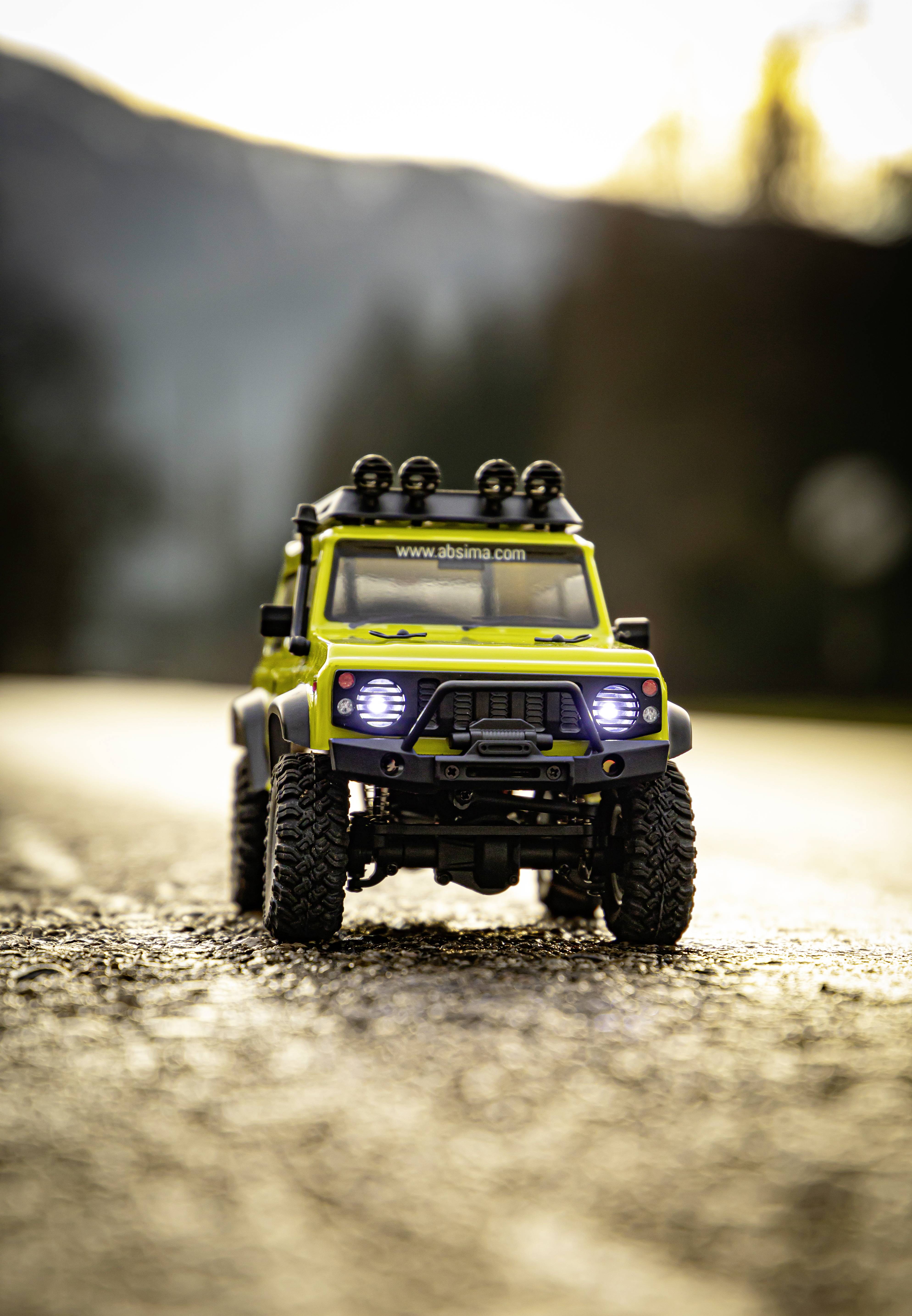 Un modèle de véhicule tout-terrain télécommandé jaune roule sur une route de campagne au coucher du soleil. Des arbres et des montagnes sont visibles en arrière-plan.