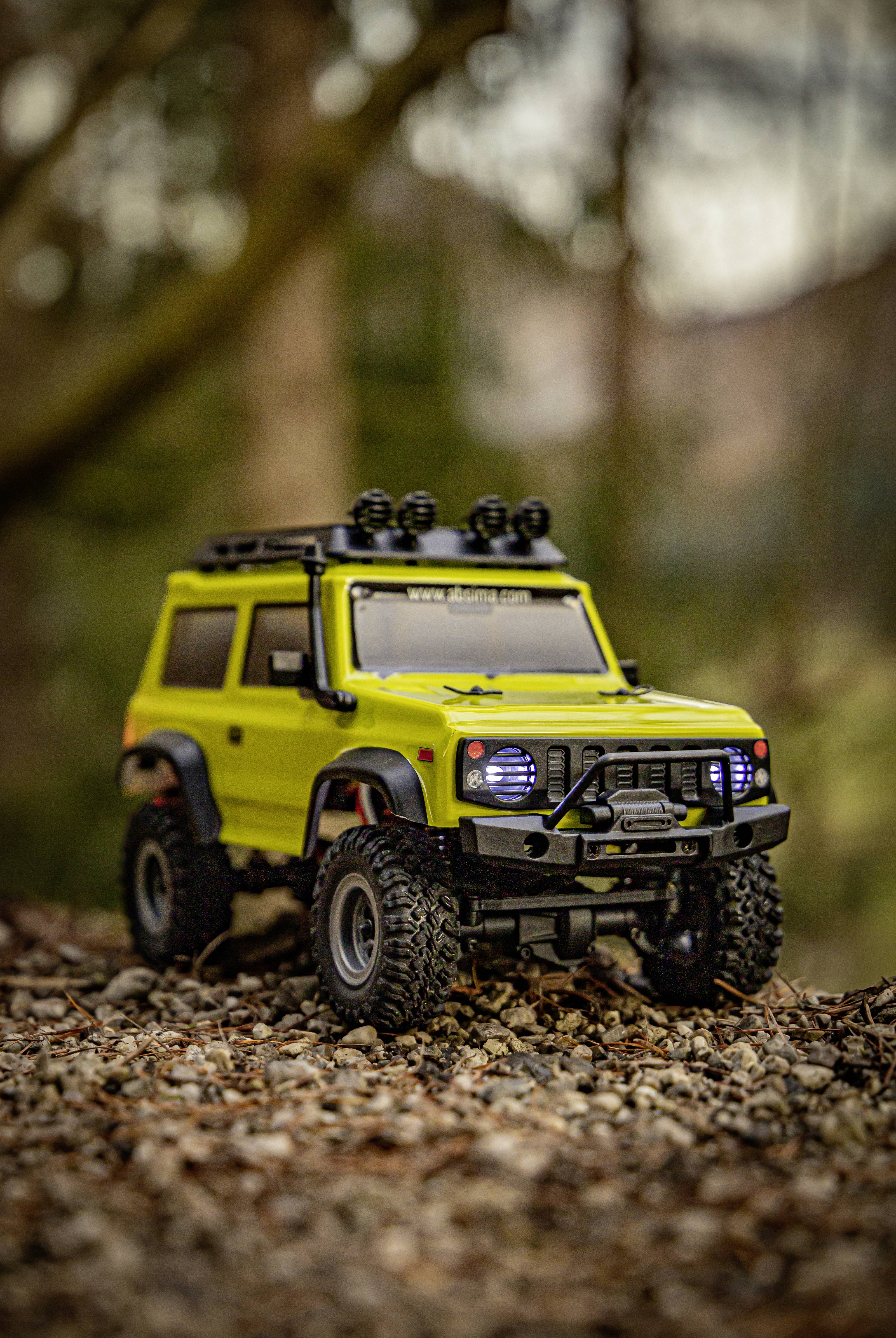 Une petite voiture miniature ressemblant à un véhicule tout-terrain vert, posée sur un sol caillouteux avec une forêt en arrière-plan.