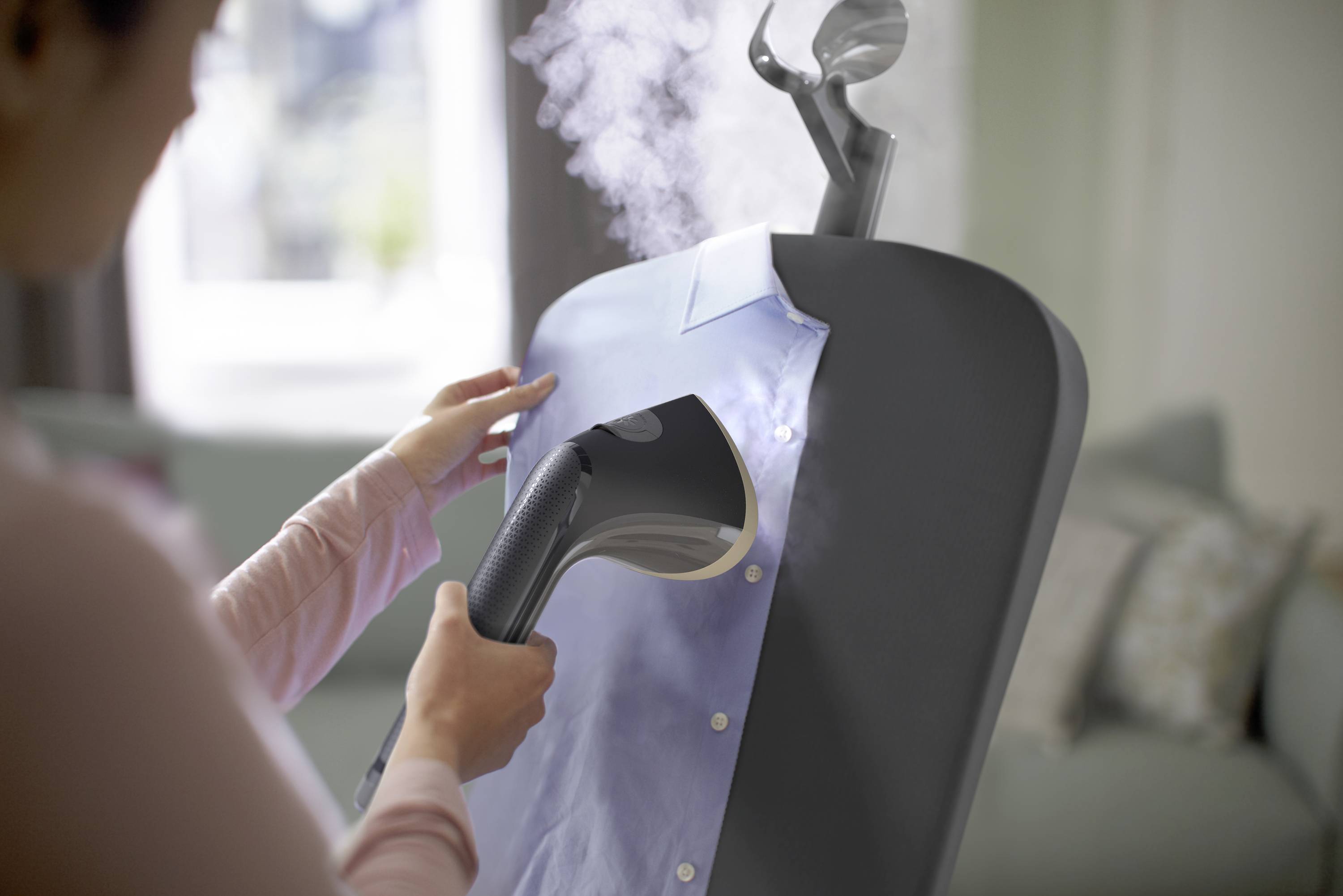 'Une personne utilise un fer à vapeur. De la vapeur est soufflée sur une chemise pour enlever les plis. L'arrière-plan flou suggère un intérieur.'