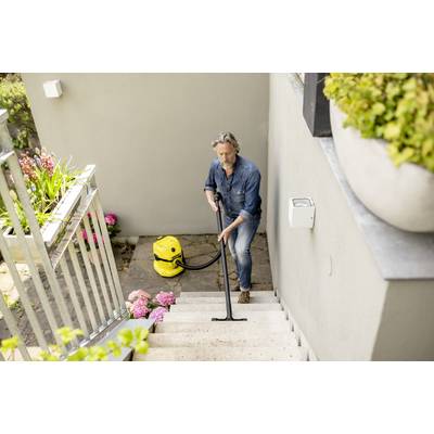 Un homme nettoie un escalier à l'extérieur avec un aspirateur jaune. À côté de lui, des hortensias roses et des plantes vertes sont en fleur.