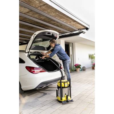 Un homme décharge le coffre d'une voiture blanche. Un aspirateur jaune est posé à côté, sous un auvent. Des pots de fleurs sont visibles en arrière-plan.