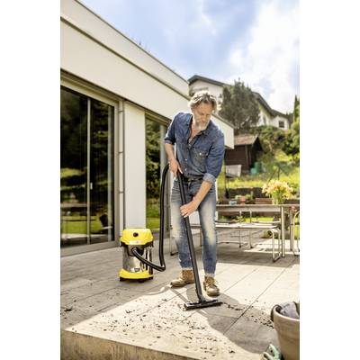 Un homme nettoie une terrasse avec un aspirateur jaune. À l'arrière-plan, on aperçoit une maison moderne et un paysage verdoyant.