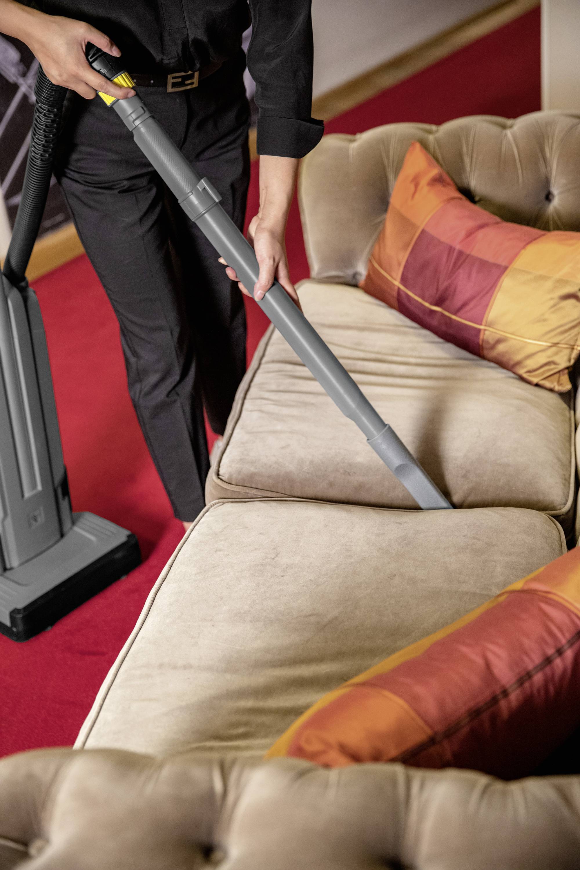 Une personne aspire un canapé de couleur beige avec un aspirateur, avec des coussins à carreaux orange et rouge visibles.