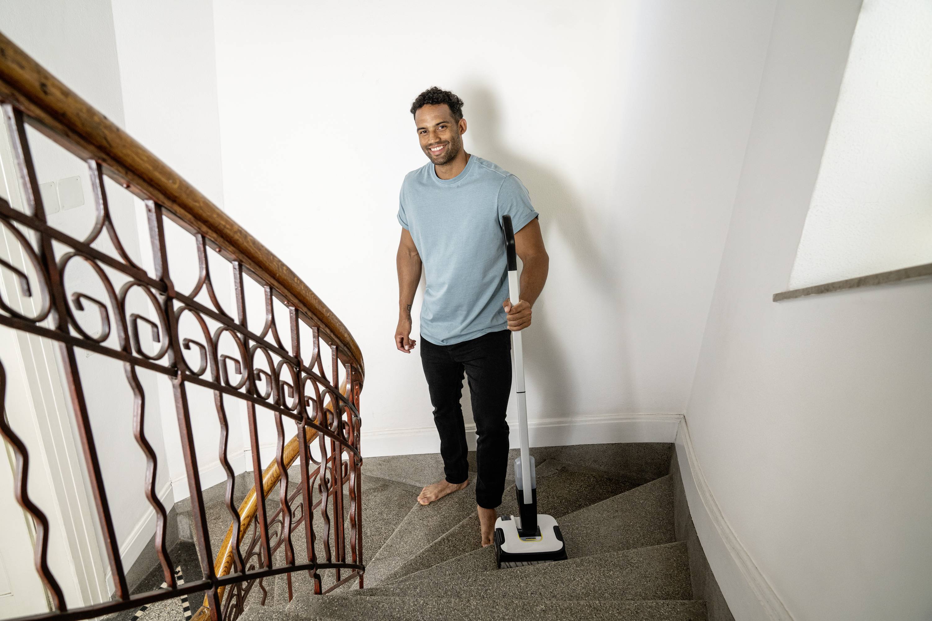 Un homme se tient pieds nus sur un escalier et tient un aspirateur. Il sourit et semble être en train de nettoyer.